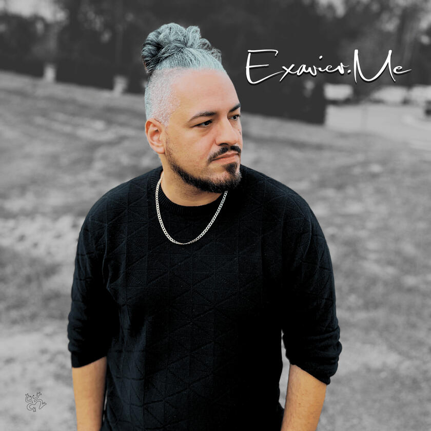 Exavier Me standing and he looks off to the side, wearing a black sweater shirt and his man-bun very present in the image.