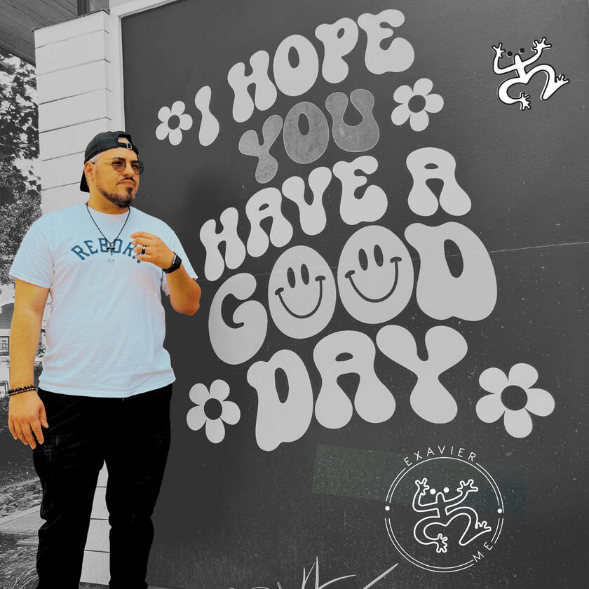Exavier Me standing in front of a sign that reads "I hope you have a good day" while wearing a backwards snap-back cap, circular sun glasses and a T-Shirt that reads "Reborn."