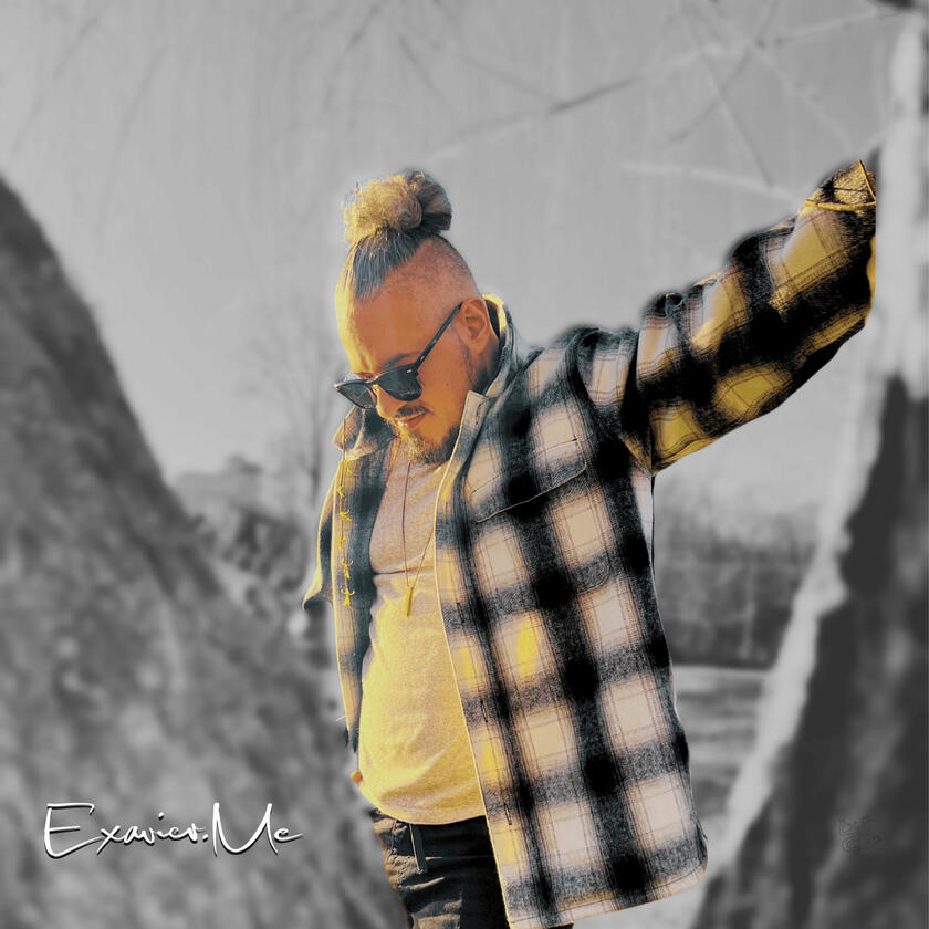 Exavier Me posing behind a tree, with a left hand leaning on a tree, the other in his pocket, as he looks down, his man bun and sunglasses are very noticeable.